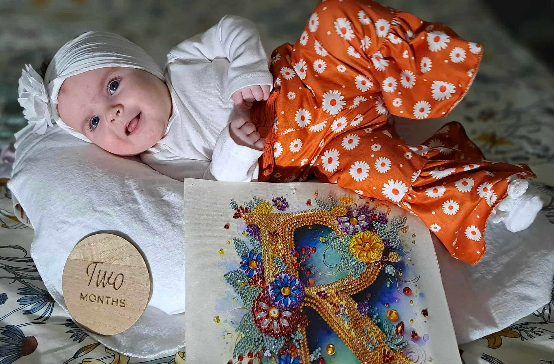 Ryleiah is registered to the contest to win money with this photo: baby, bejeweled_letter, child, closeup, colorful_art, cute, floral_pattern, flower, happy, headband, indoors, infant, lying_down, orange_pants, pillow, portrait, smiling, two_months, white_top, wooden_plaque