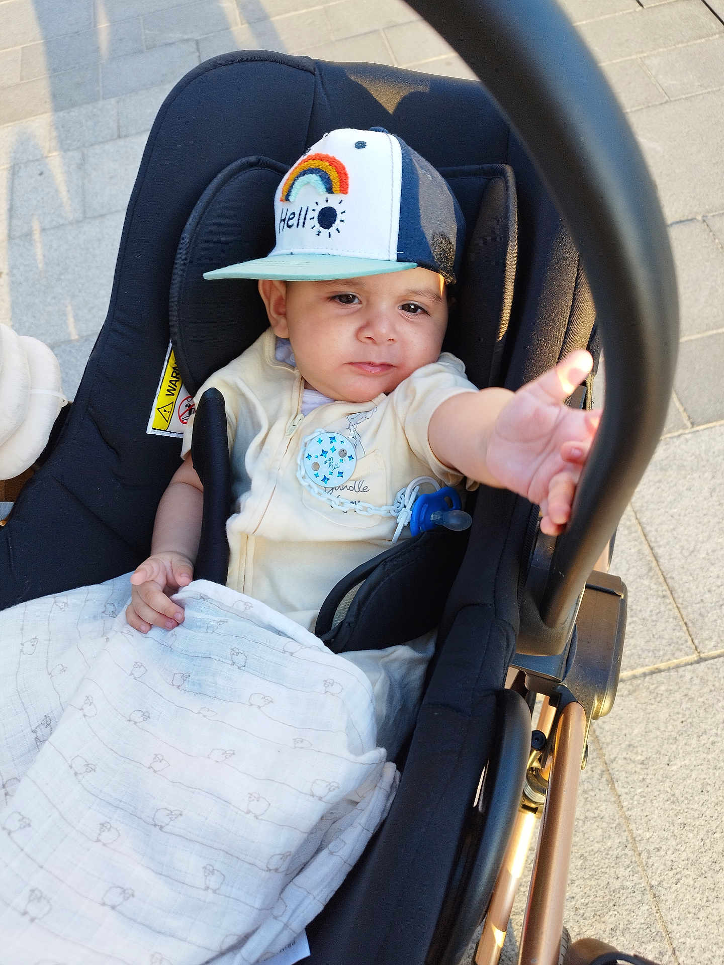Aryane a rejoint le concours — aidez-le/la à gagner de superbes lots ! baby, blanket, cap, casual, child, clothing, comfort, curious, cute, hand, infant, outdoor, pavement, person, reaching, seat, stroller, sunlight, transport, young