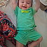 baby, child, smile, green_clothing, shorts, tshirt, bed, bedding, patterned_fabric, happy, arms_up, lying_down, infant, person, indoors, cute, joyful, face, young_child, playful