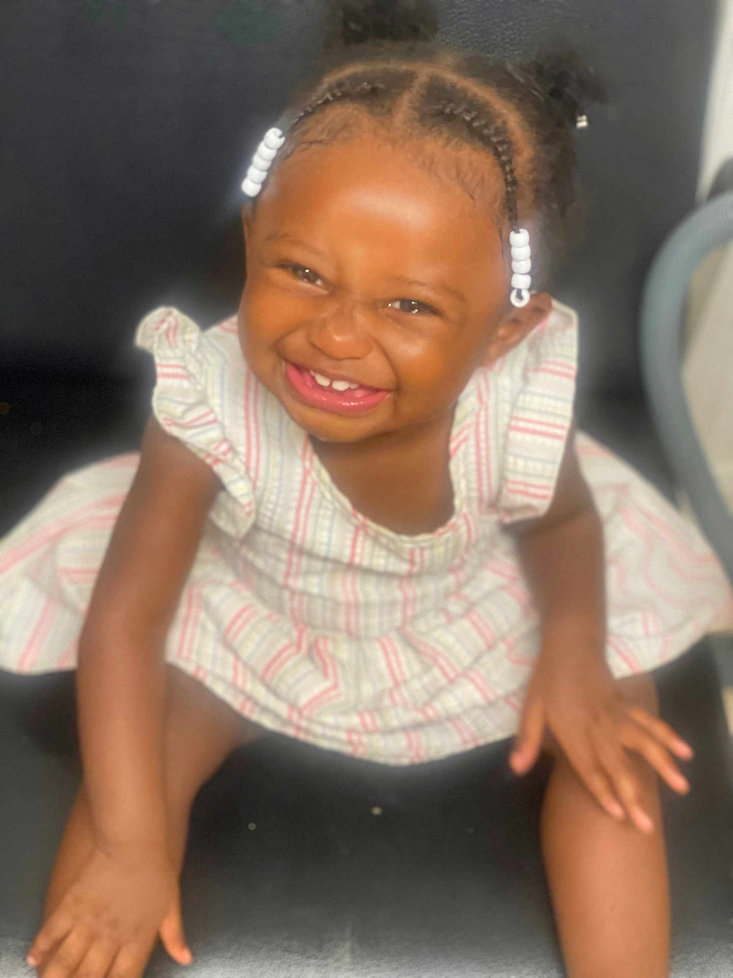 Dionasia is registered to the contest to win money with this photo: toddler, child, smiling, braids, beads, striped_dress, sitting, black_chair, happy, cute, portrait, indoors, skin, hair, young_child, person, face, laughing, baby, fun