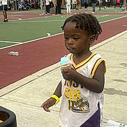Lamont is registered to the contest to win money with this photo: toddler, child, ice_cream, basketball_court, outdoor, sport_clothing, yellow_shorts, white_jersey, people, playing, summer, daytime, sidewalk, trees, tent, recreation, fun, casual, person, event
