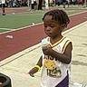 toddler, child, ice_cream, basketball_court, outdoor, sport_clothing, yellow_shorts, white_jersey, people, playing, summer, daytime, sidewalk, trees, tent, recreation, fun, casual, person, event