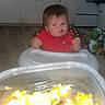 toddler, child, high_chair, red_shirt, food_container, fork, kitchen, floor, oven, cabinet, toy, yoshi, person, indoor, expression, furniture, baby, curious, sitting, meal