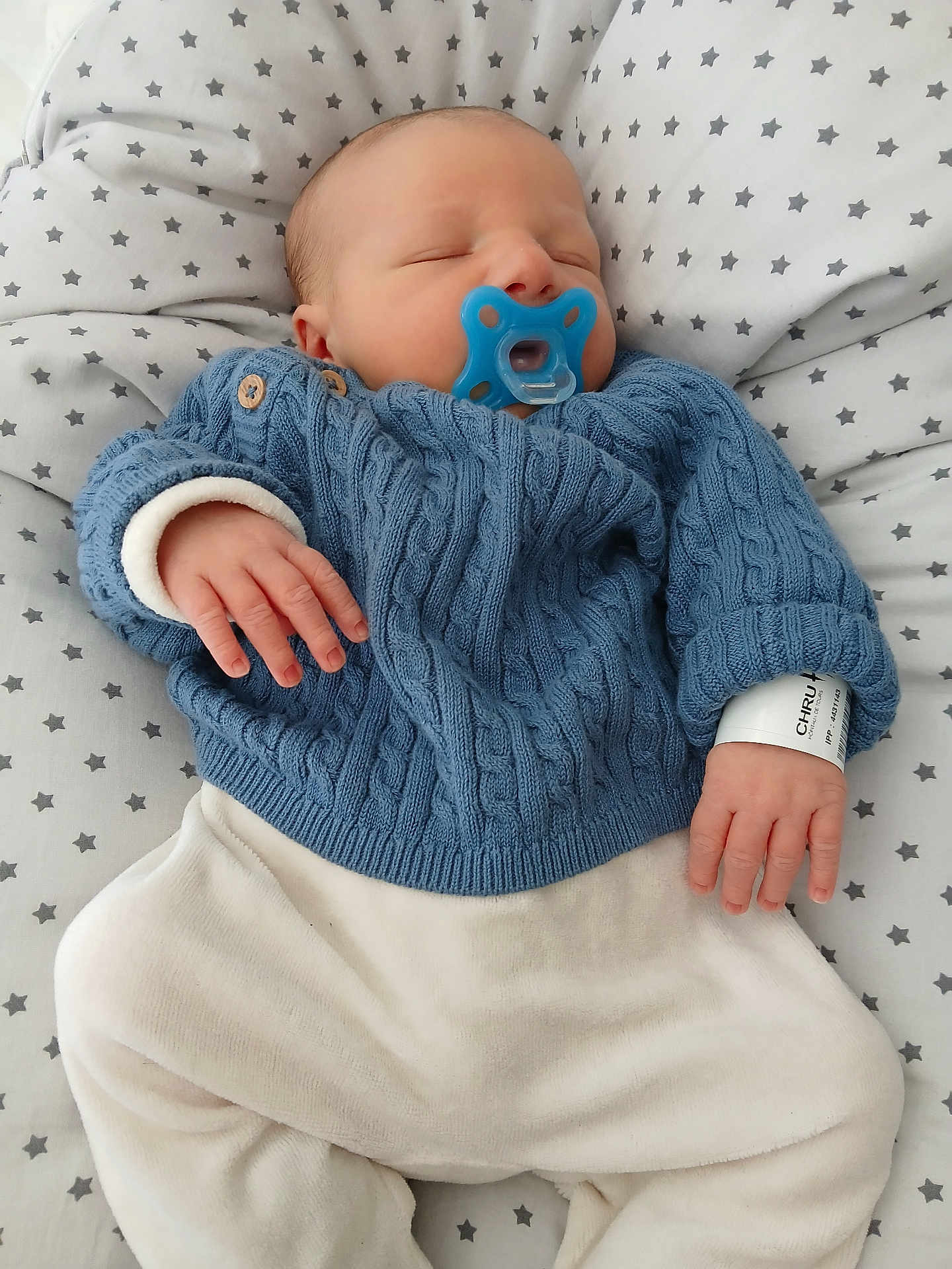 Sohan participe au concours pour gagner de l'argent avec cette photo : baby, newborn, sleeping, pacifier, blue_sweater, white_pants, hospital_wristband, star_pattern, blanket, infant, cozy, cute, child, resting, hand, face, soft, indoors, clothing, portrait