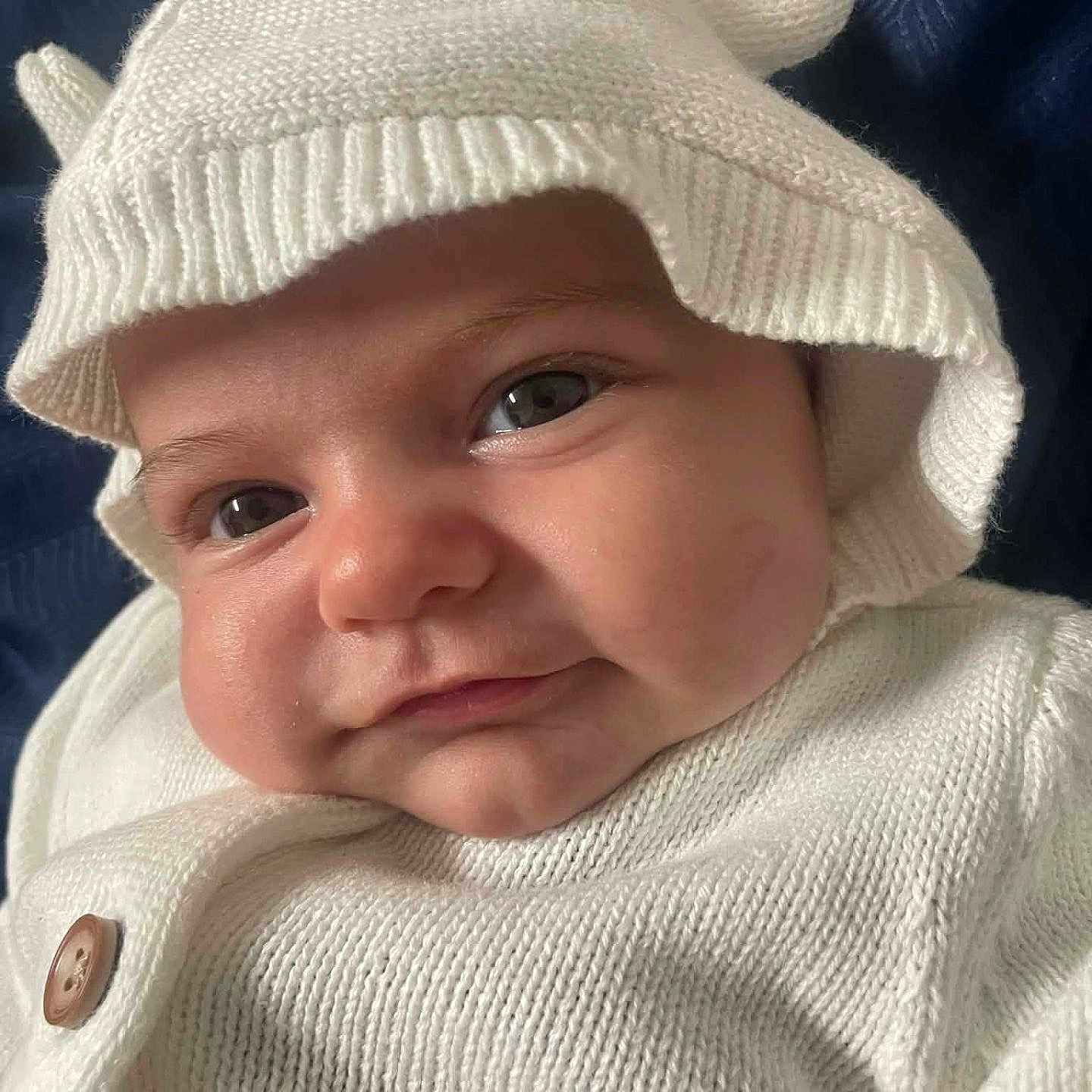 Makinsey is registered to the contest to win money with this photo: baby, baby_face, blue_background, button, cheeks, child, close_up, cute, eyes, face, hat, headwear, indoors, infant, knitwear, portrait, smile, soft_texture, warm_clothing, white_clothing