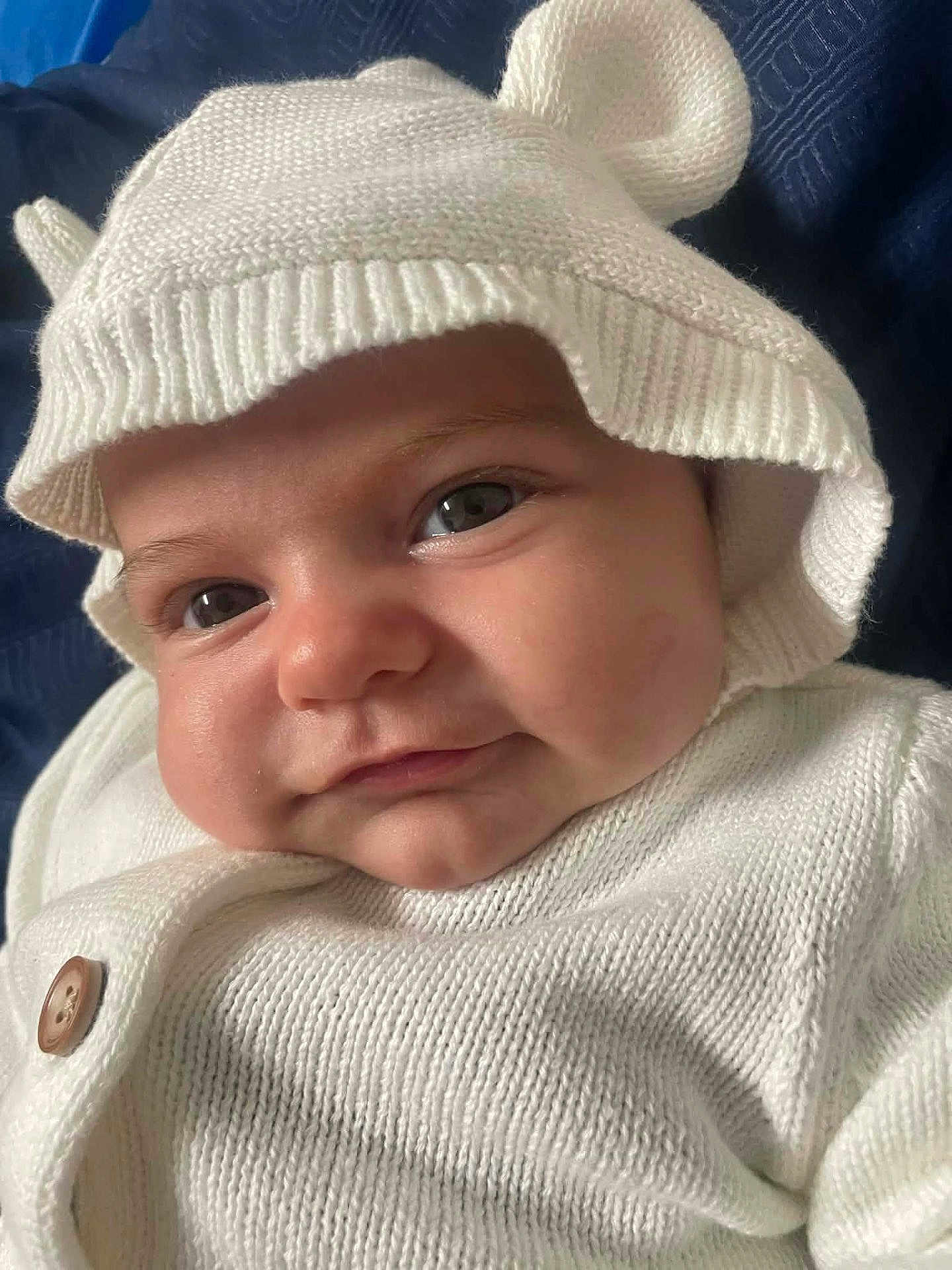 Makinsey is registered to the contest to win money with this photo: baby, infant, face, hat, knitwear, white_clothing, cute, smile, close_up, child, portrait, baby_face, warm_clothing, button, headwear, soft_texture, indoors, blue_background, eyes, cheeks