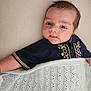 baby, infant, blanket, blue_shirt, embroidered_clothing, face, skin, cute, lying_down, indoors, soft_texture, arm, head, expression, young_child, portrait, cozy, warm, fabric, resting