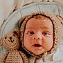 baby, infant, knitted_hat, bear_ears, crocheted_teddy_bear, blanket, cozy, cute, wide_eyes, surprised_expression, soft_texture, portrait, child, newborn, indoors, warm, snuggled, sleeping_area, adorable, headwear