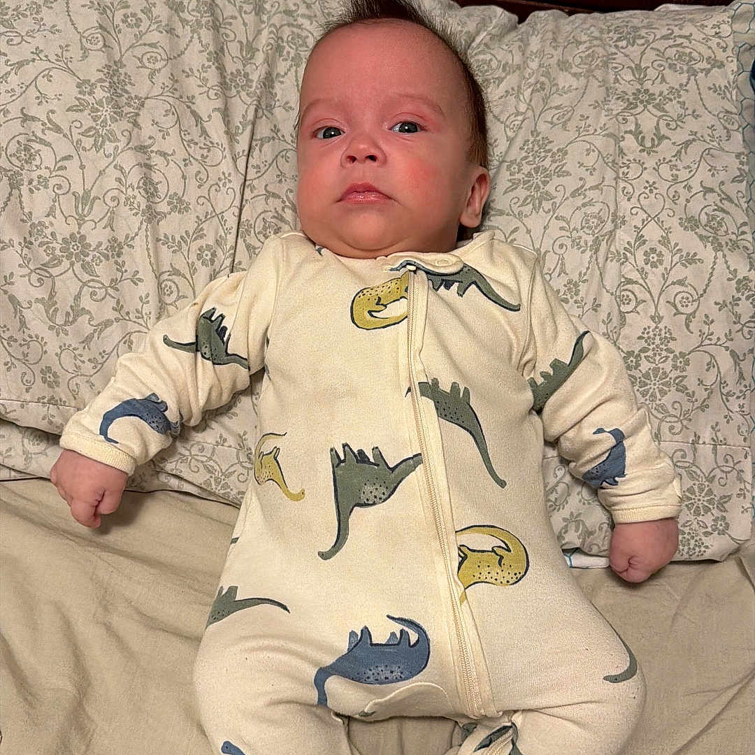 Alejandro Guerrero joined the competition — help win amazing prizes! baby, bed, bedding, child, cute, dinosaur_print, expression, fabric, face, indoors, infant, lying_down, newborn, onesie, person, pillow, portrait, sleepwear, socks, wooden_headboard