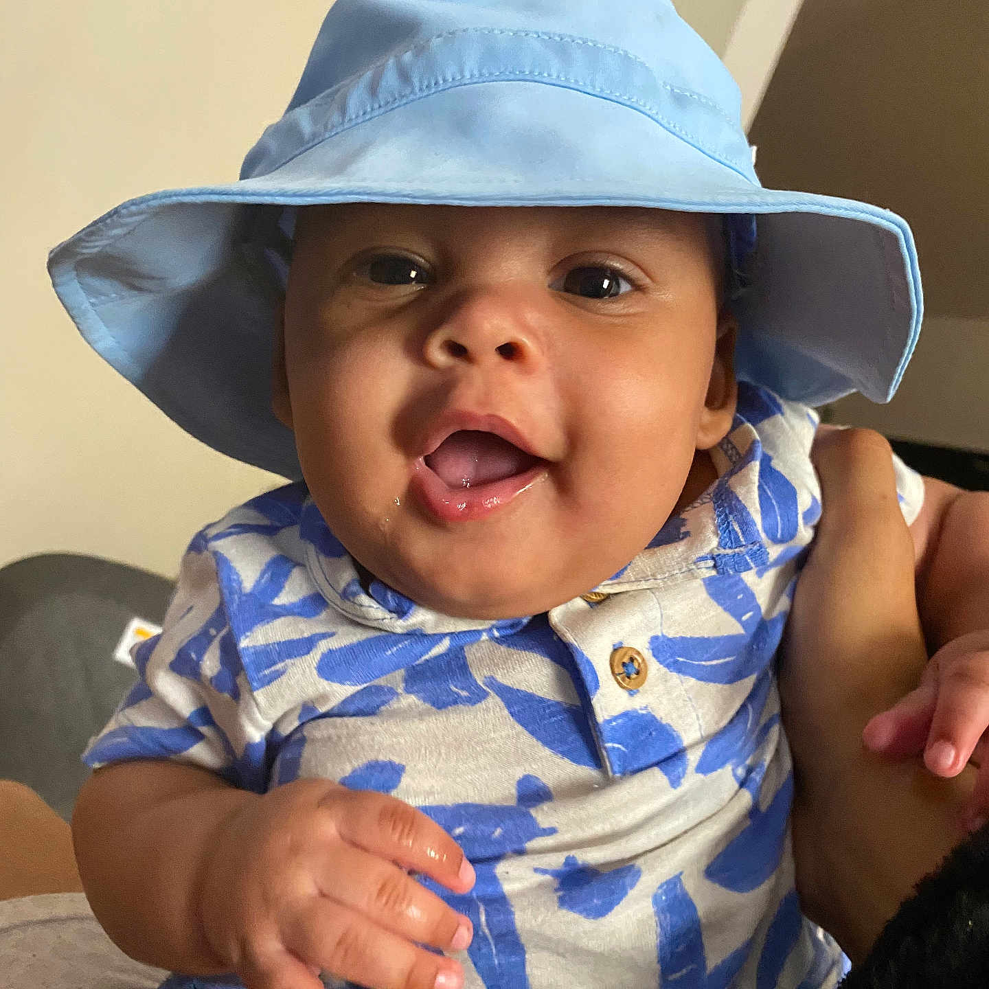 Aramis is registered to the contest to win money with this photo: baby, blue_hat, child, clothing, cute, expression, face, hand, happy, hat, holding, indoor, infant, patterned_shirt, person, portrait, sitting, skin, smiling, young