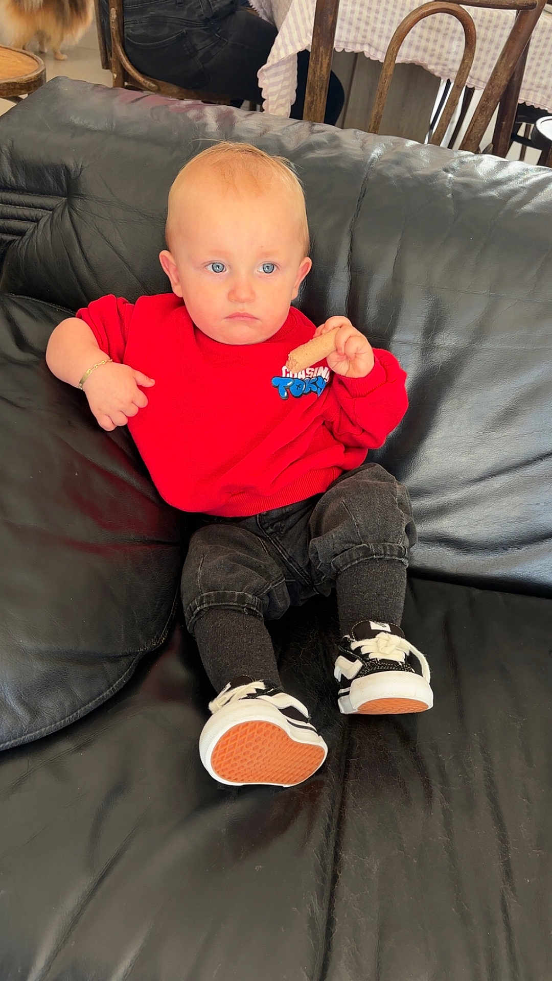 Knay Kraemer a rejoint le concours — aidez-le/la à gagner de superbes lots ! toddler, child, red_sweatshirt, black_pants, sneakers, black_leather_couch, snack, indoor, person, seated, blue_eyes, short_hair, casual_clothing, bracelet, table, chair, floor, background_person, home, relaxed