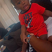 Christian is registered to the contest to win money with this photo: child, toddler, baby, portrait, sitting, indoors, red_clothing, smiling, curious, watch, smartwatch, adult, lap, sofa, cushions, window_blinds, home, black_socks, toy, photograph
