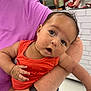 baby, infant, child, person, holding, orange_clothing, purple_clothing, arm, watch, indoor, store, people, blurred_background, curious, expression, head_tilt, skin, face, eyes, mouth