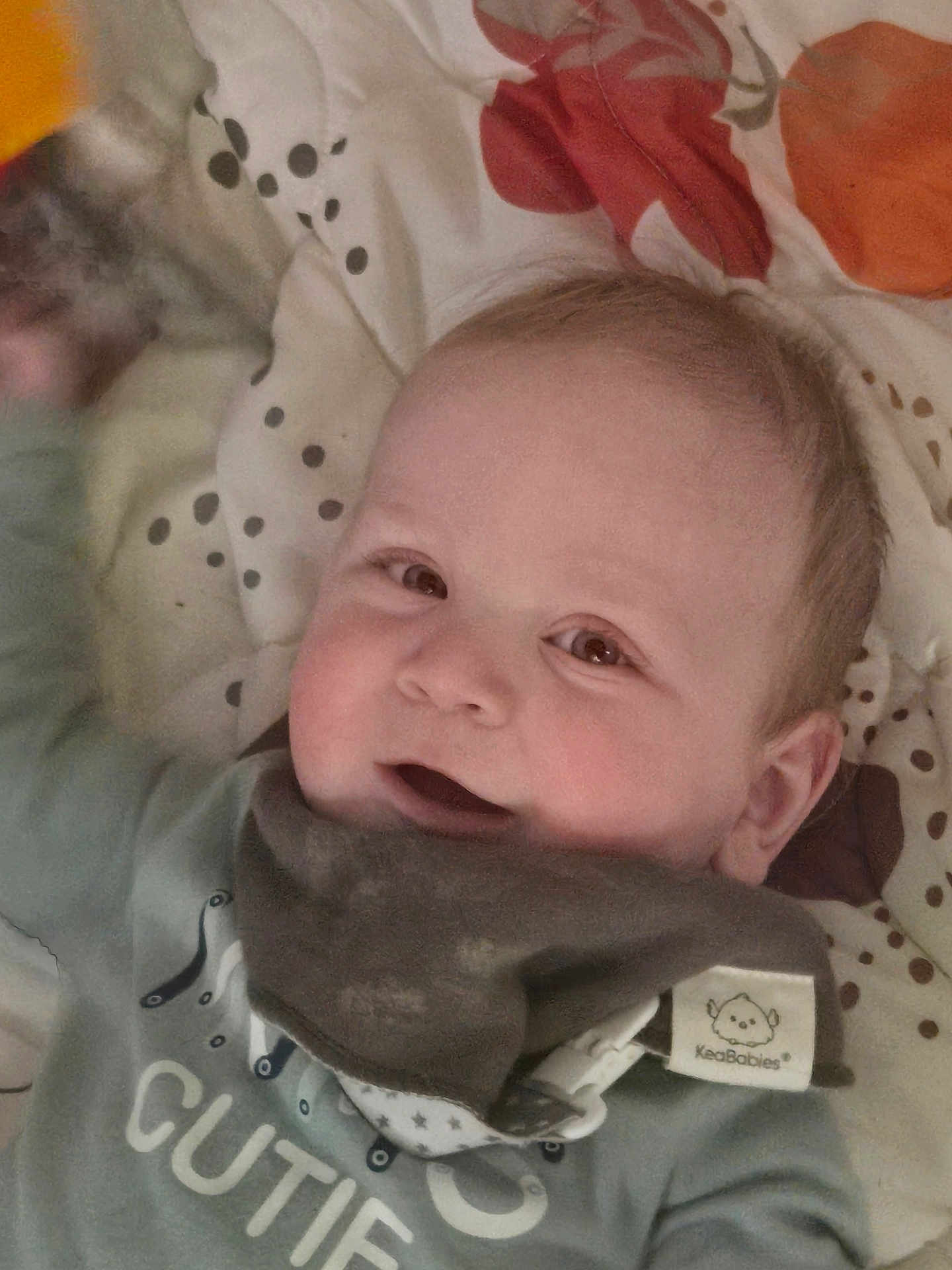 Ezra joined the competition — help win amazing prizes! baby, infant, child, smile, cheeks, eyes, face, bib, blanket, polka_dot, clothing, head, ear, mouth, lying_down, bed, cute, adorable, portrait, home