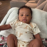 Makai is registered to the contest to win money with this photo: baby, infant, child, onesie, giraffe_pattern, blanket, curly_hair, living_room, couch, soft_toy, floor, wood_floor, person, cute, relaxed, indoors, home, comfort, portrait, young