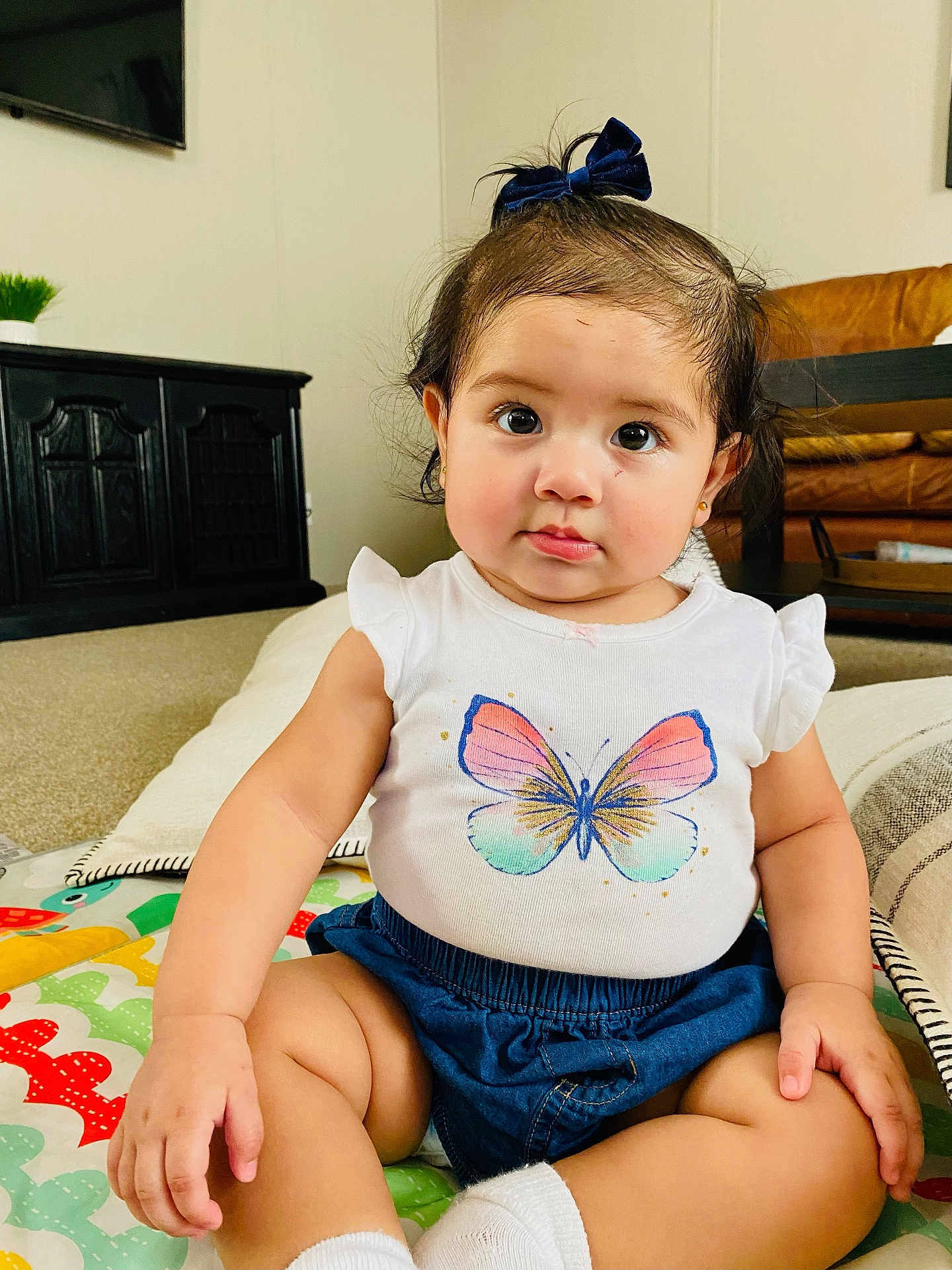 Emma joined the competition — help win amazing prizes! baby, child, infant, sitting, indoor, play_mat, butterfly_print, white_shirt, denim_shorts, blue_bow, cute, curious_eyes, chubby_cheeks, living_room, couch, carpet, pillow, furniture, person, portrait