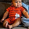 baby, child, infant, pouty_face, orange_clothing, striped_clothing, elephant_patch, couch, gray_fabric, blanket, sitting, indoors, cute, expression, barefoot, chubby_cheeks, short_hair, relaxed, home, comfort
