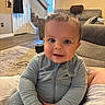 Kamari is registered to the contest to win money with this photo: baby, child, indoor, living_room, couch, stairs, clothing, onesie, portrait, smile, face, person, home, furniture, photography, curious, cozy, wall_art, door, carpet