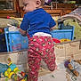child, toddler, standing, blue_shirt, monkey_print_pants, toy, playpen, indoor, carpet, stuffed_animal, baby_toy, colorful, clothing, furniture, person, home, floor_mat, plush_toy, crawling_area, cute