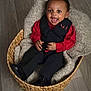 baby, toddler, smiling, basket, blanket, clothing, red_shirt, black_vest, black_pants, footwear, floor, happy, portrait, indoors, child, cute, sitting, expression, person, cozy