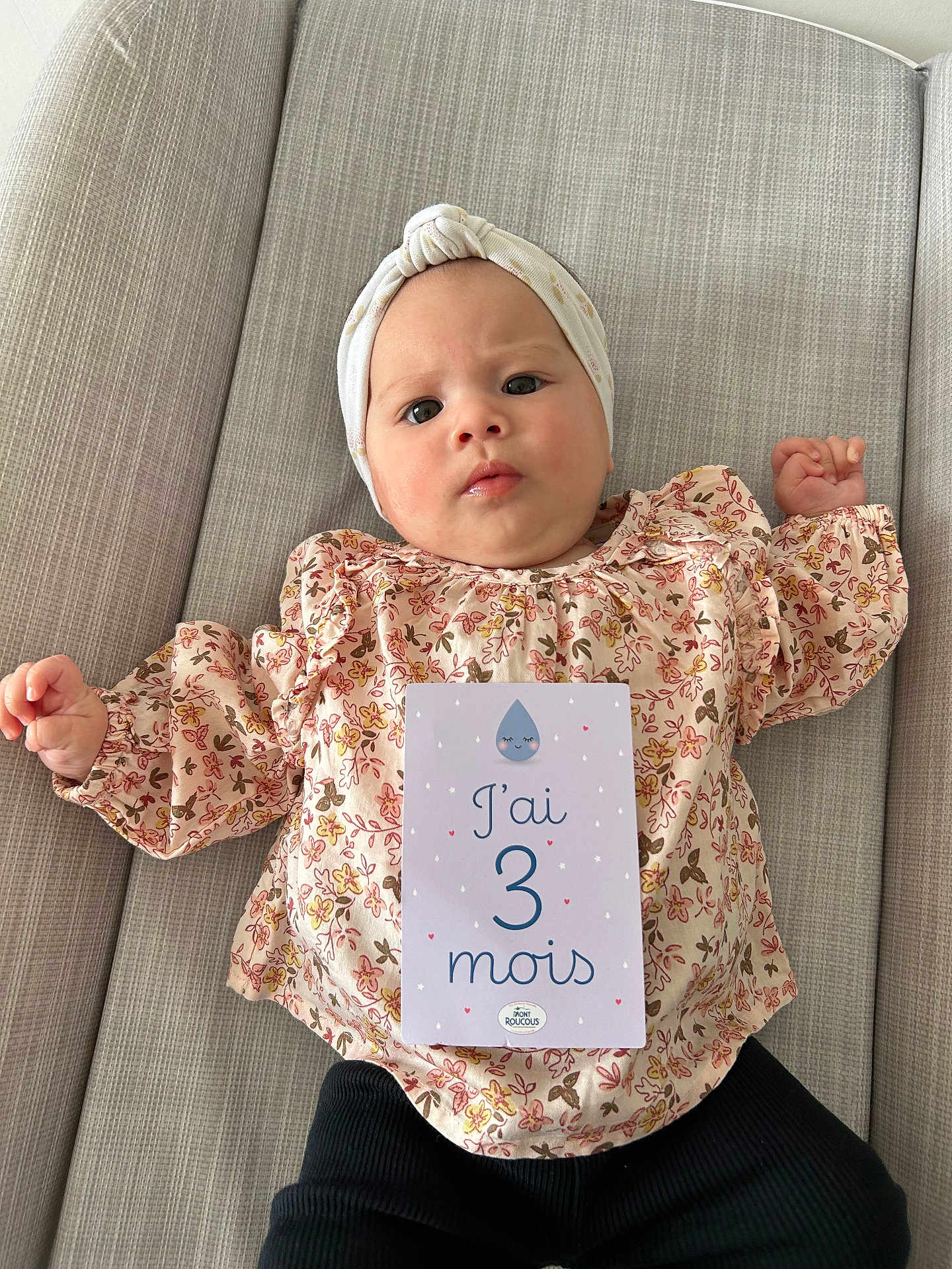 Esmée participe au concours pour gagner de l'argent avec cette photo : baby, infant, milestone_card, floral_clothing, head_wrap, couch, gray_couch, milestone, card, person, child, indoors, cute, portrait, laying_down, expressive_face, pink, black_pants, fabric, home