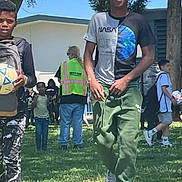 Antoine joined the competition — help win amazing prizes! boy, child, teen, soccer_ball, grass, outdoor, sunny_day, people, walking, safety_vest, building, tree, casual_clothing, smiling, group, shoes, shorts, tshirt, green_pants, recess