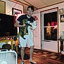 man, cat, indoor, barefoot, shorts, tshirt, brown_tabby_cat, white_cat, wooden_floor, television, rubber_ducks, door, window, fan, table, rug, lamp, picture_frame, socks, living_room