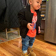 Bronx is registered to the contest to win money with this photo: toddler, child, boots, jacket, jeans, orange_shirt, standing, indoor, wooden_floor, earrings, curly_hair, clothing, person, flooring, casual_wear, young_child, furniture, home_interior, light_bokeh, expression