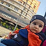 Léo participe au concours pour gagner de l'argent avec cette photo : child, toddler, baby_carrier, orange_jacket, hat, outdoor, urban, building, sky, person, face, eyes, hand, clothing, portrait, casual, daylight, expression, seating, blurred_background