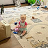 toddler, child, toy, rug, living_room, couch, blanket, floor, dinosaur_toy, pink_cup, blue_fish_toy, green_dinosaur, walker, wooden_floor, playtime, indoor, person, sitting, curly_hair, cute