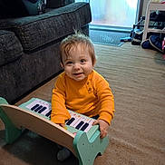 Maddox is registered to the contest to win money with this photo: baby, child, toy_piano, orange_sweater, carpet, indoor, living_room, smiling, playing, furniture, window, door, couch, person, happy, floor, head, hair, hand, seat