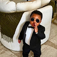 Israel is registered to the contest to win money with this photo: toddler, child, sunglasses, tuxedo, bow_tie, indoor, carpet, couch, blanket, person, fashion, portrait, standing, curly_hair, clothing, formal_wear, cute, confident, home, furniture