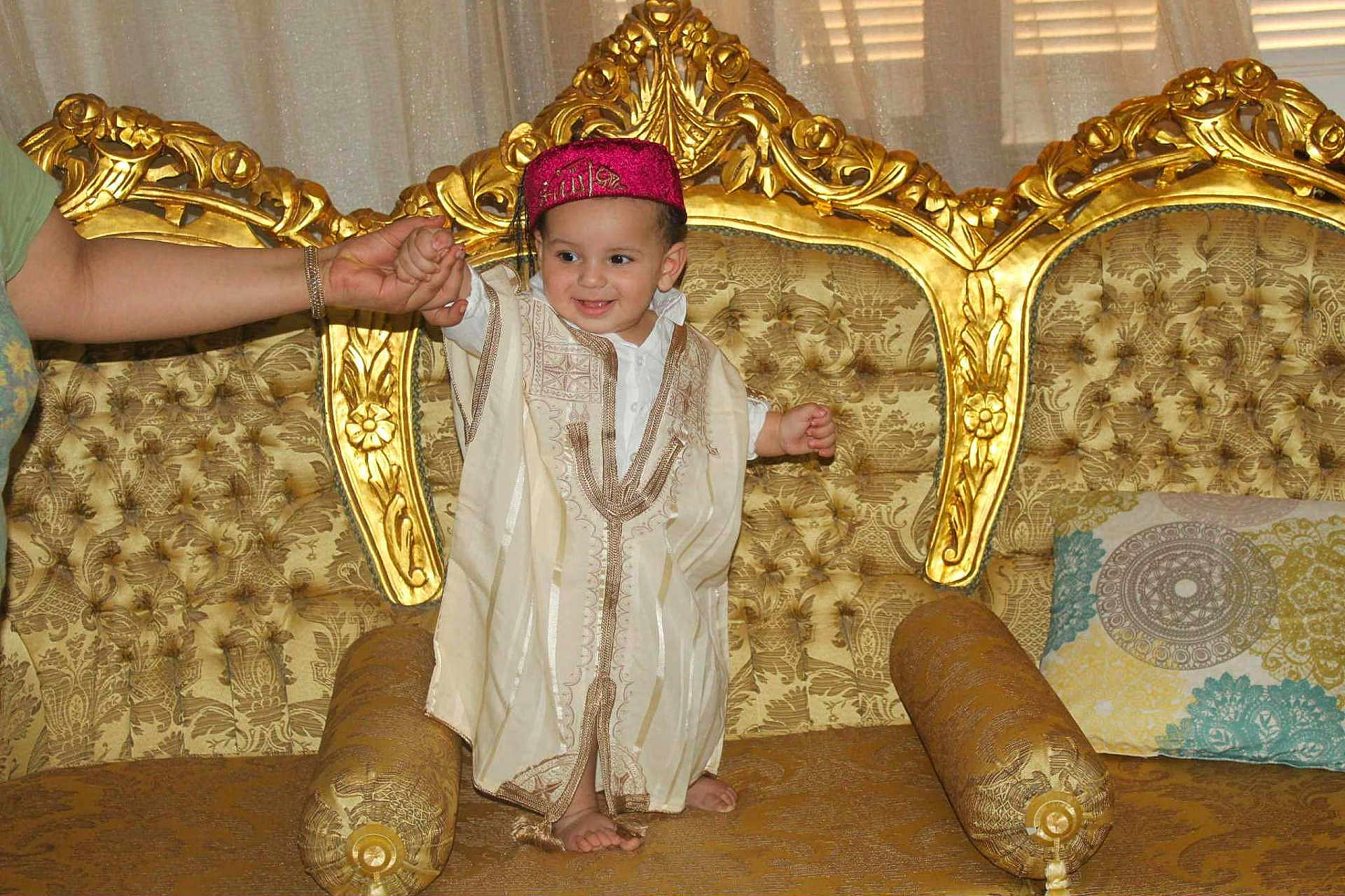 Soulayman participe au concours pour gagner de l'argent avec cette photo : toddler, child, traditional_clothing, sofa, golden, ornate, couch, smiling, hand_holding, person, indoor, home, furniture, decor, pillow, footwearless, curtain, happy, baby, portrait