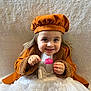 child, toddler, smile, face, beret, coat, brown_coat, white_dress, lollipop, happy, cute, portrait, sitting, indoor, soft_background, long_hair, hands, playful, fashion, warm_clothing