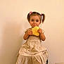 beige_dress, bread, child, cushion, cute, food, girl, hair_clips, happy, indoor, light_skin, pigtails, playful, portrait, sitting, smile, snack, toddler, white_sneakers, young_child