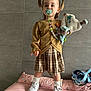 brown_cardigan, child, clothing, cute, floor_cushion, indoor, minnie_mouse, pacifier, person, pigtails, pink_cushion, plaid_skirt, plush_toy, smiling, standing, toddler, toy, wall_tiles, white_sneakers, white_socks