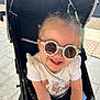 accessory, baby, casual_clothing, child, cute, daytime, fashion, hair, happy, outdoor, person, playful, seatbelt, shorts, smile, stroller, summer, sunglasses, sunlight, toddler