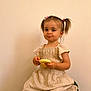 beige_wall, bread, calm, casual, child, cute, dress, food, girl, hair_accessories, holding, indoors, neutral_background, person, pigtails, portrait, sitting, small_child, smile, young_child