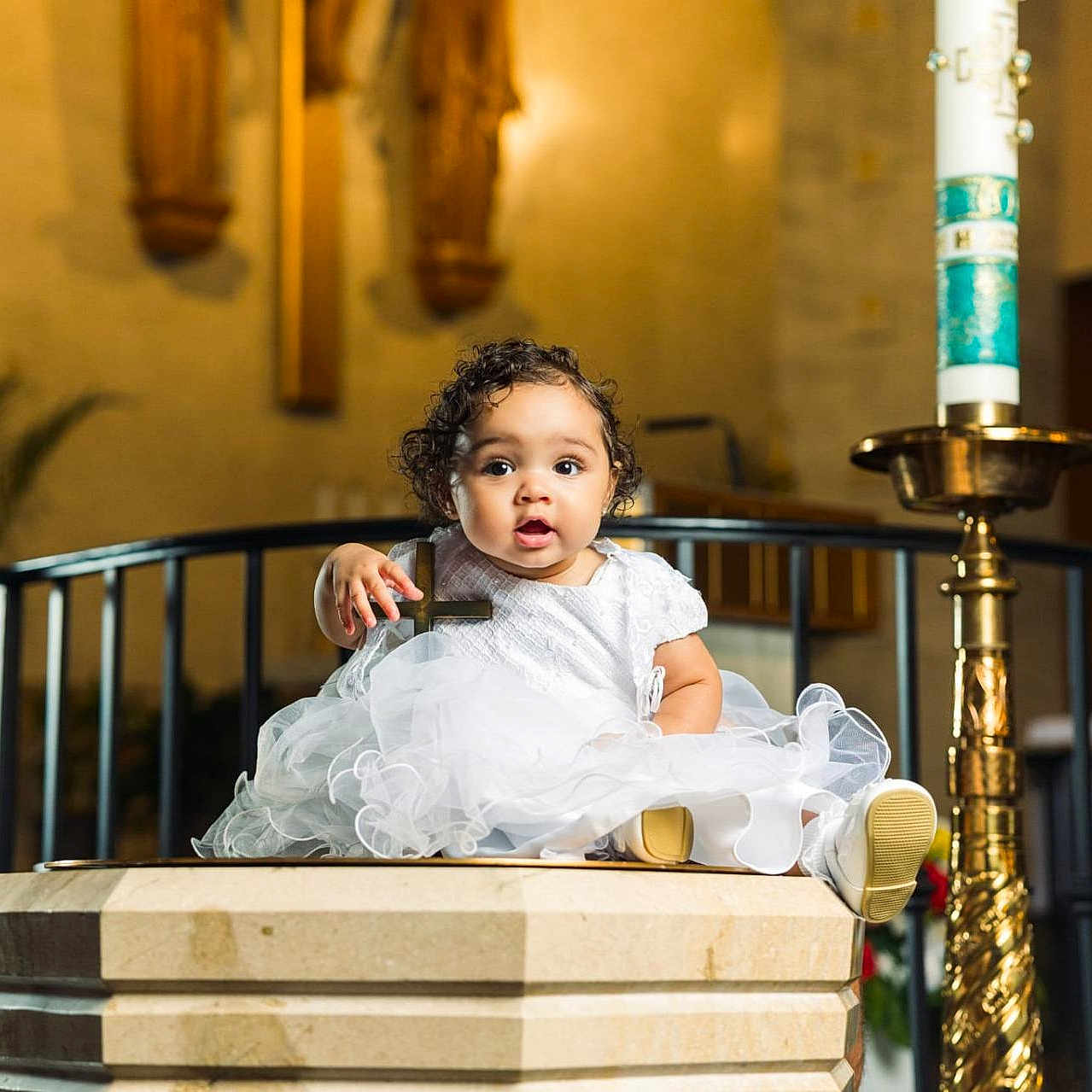 Emmi joined the competition — help win amazing prizes! baby, child, infant, portrait, church, baptism, baptismal_font, white_dress, formal_clothing, curly_hair, smiling, wide_eyed, sitting, ceremonial_candle, religious_setting, indoor, warm_lighting, stone, ornate, photography