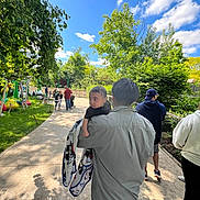 Emiliano is registered to the contest to win money with this photo: child, adult, outdoor, park, greenery, trees, sky, clouds, people, pathway, blanket, casual_clothing, walking, daylight, family, nature, sculptures, shadows, leaves, stroller