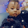 black_clothing, buttons, casual_pose, child, colorful_lighting, curious_expression, dreadlocks, face, hairstyle, head, indoor, jacket, looking_at_camera, modern_interior, person, portrait, sitting, social_media_interface, timestamp, young