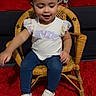 child, toddler, girl, sitting, portrait, smiling, happy, face, bows, hair_accessories, white_shirt, jeans, sneakers, rattan_chair, wicker_furniture, red_carpet, couch, indoor, fashion, photography