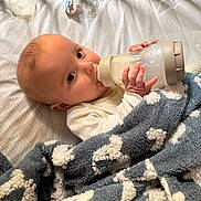 Kye is registered to the contest to win money with this photo: baby, bottle, blanket, crib, milk, infant, bed, sleeping_area, cozy, child, feeding, blanket_pattern, hand, face, person, indoors, soft_texture, pacifier, nursery, resting