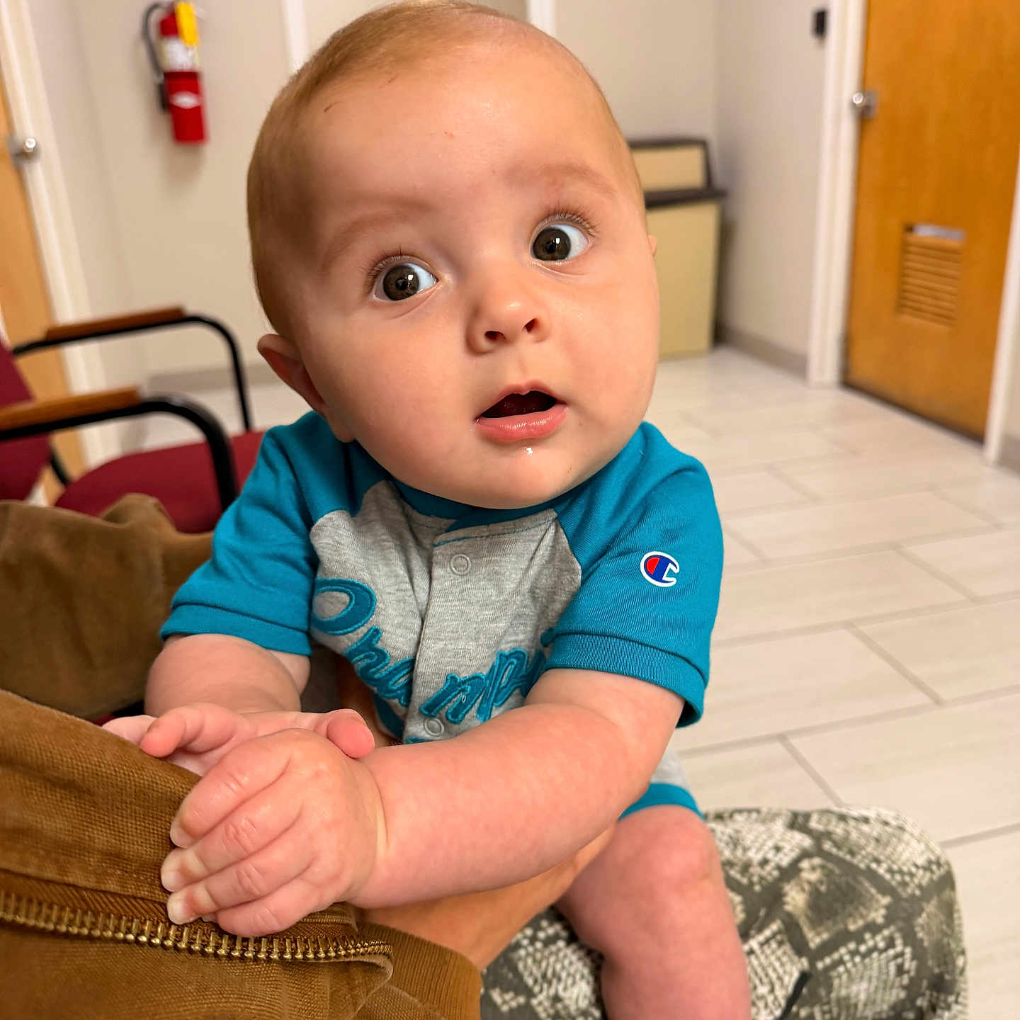 Kye is registered to the contest to win money with this photo: baby, infant, child, blue_shirt, curious_expression, indoor, person, hand, arm, face, eyes, seated, floor, door, chair, fire_extinguisher, casual_clothing, skin, young_child, grip