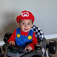 Cannon is registered to the contest to win money with this photo: toddler, child, costume, mario, toy_car, race_car, indoor, kitchen, halloween, pumpkin, decoration, smiling, happy, face, hat, red_clothing, blue_overalls, floor, wheel, light
