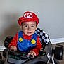 toddler, child, costume, mario, toy_car, race_car, indoor, kitchen, halloween, pumpkin, decoration, smiling, happy, face, hat, red_clothing, blue_overalls, floor, wheel, light