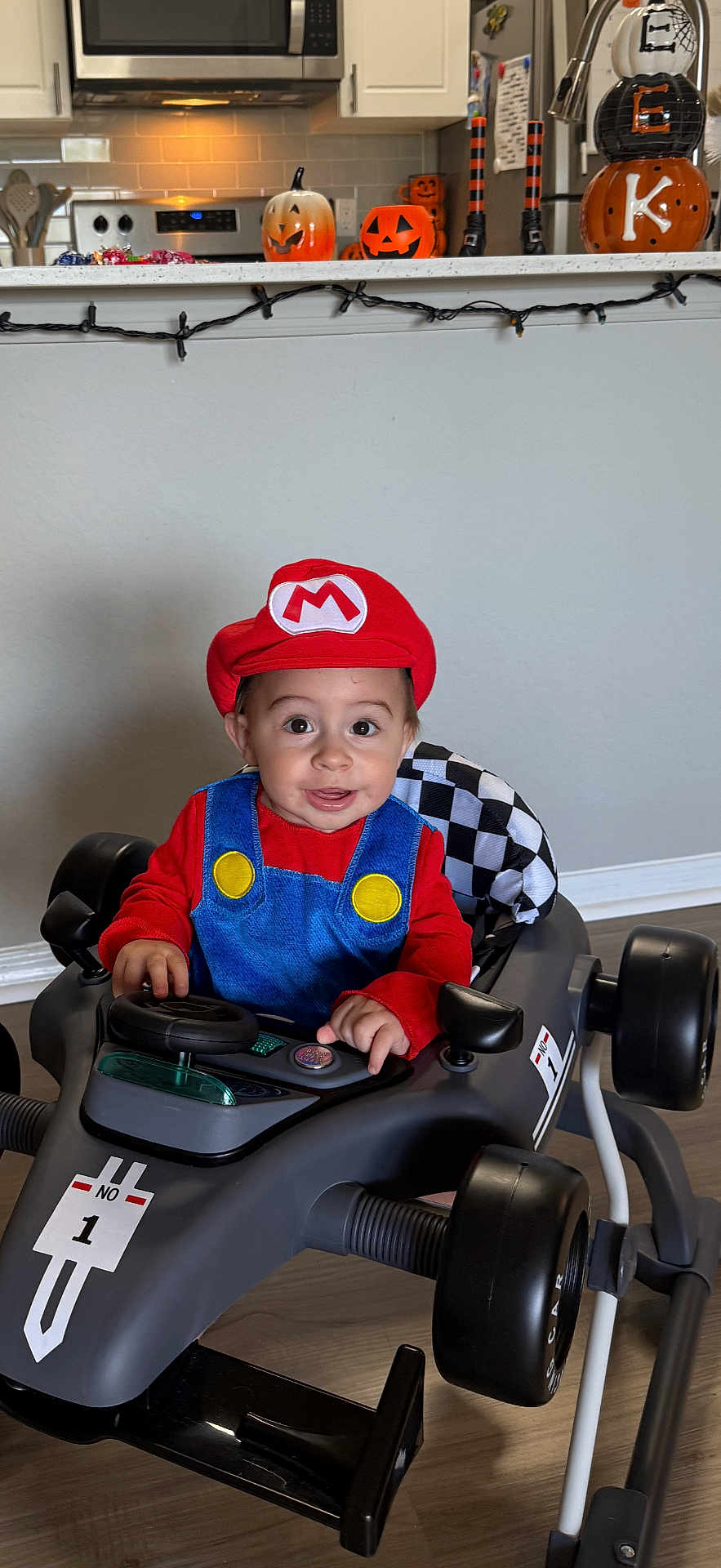 Cannon is registered to the contest to win money with this photo: toddler, child, costume, mario, toy_car, race_car, indoor, kitchen, halloween, pumpkin, decoration, smiling, happy, face, hat, red_clothing, blue_overalls, floor, wheel, light