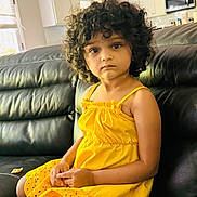 Navya joined the competition — help win amazing prizes! black_leather_couch, cabinet, casual, child, contemplative, curly_hair, expressive_eyes, furniture, home, indoor, kitchen, microwave, neutral_colors, person, portrait, relaxed, sitting, window, yellow_dress, young_child