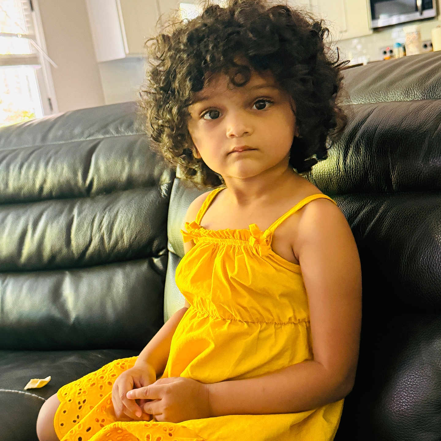 Navya joined the competition — help win amazing prizes! black_leather_couch, cabinet, casual, child, contemplative, curly_hair, expressive_eyes, furniture, home, indoor, kitchen, microwave, neutral_colors, person, portrait, relaxed, sitting, window, yellow_dress, young_child