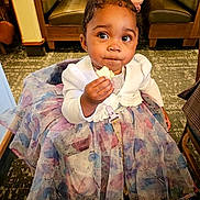 Vision is registered to the contest to win money with this photo: baby, booth, butterfly_pattern, carpet, chair, child, clothing, curious, curly_hair, cute, dress, girl, indoor, pigtails, portrait, restaurant, seated, snack, table, toddler